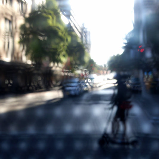 Urban Commute - Series No.2: 9230v1a - Close Detail View, fine art photograph by Raymond Mather featuring a silhouetted scooter rider crossing a sunlit city street. Seen through a patterned screen, the image blends light, blur, and architectural rhythm into a poetic urban abstraction.