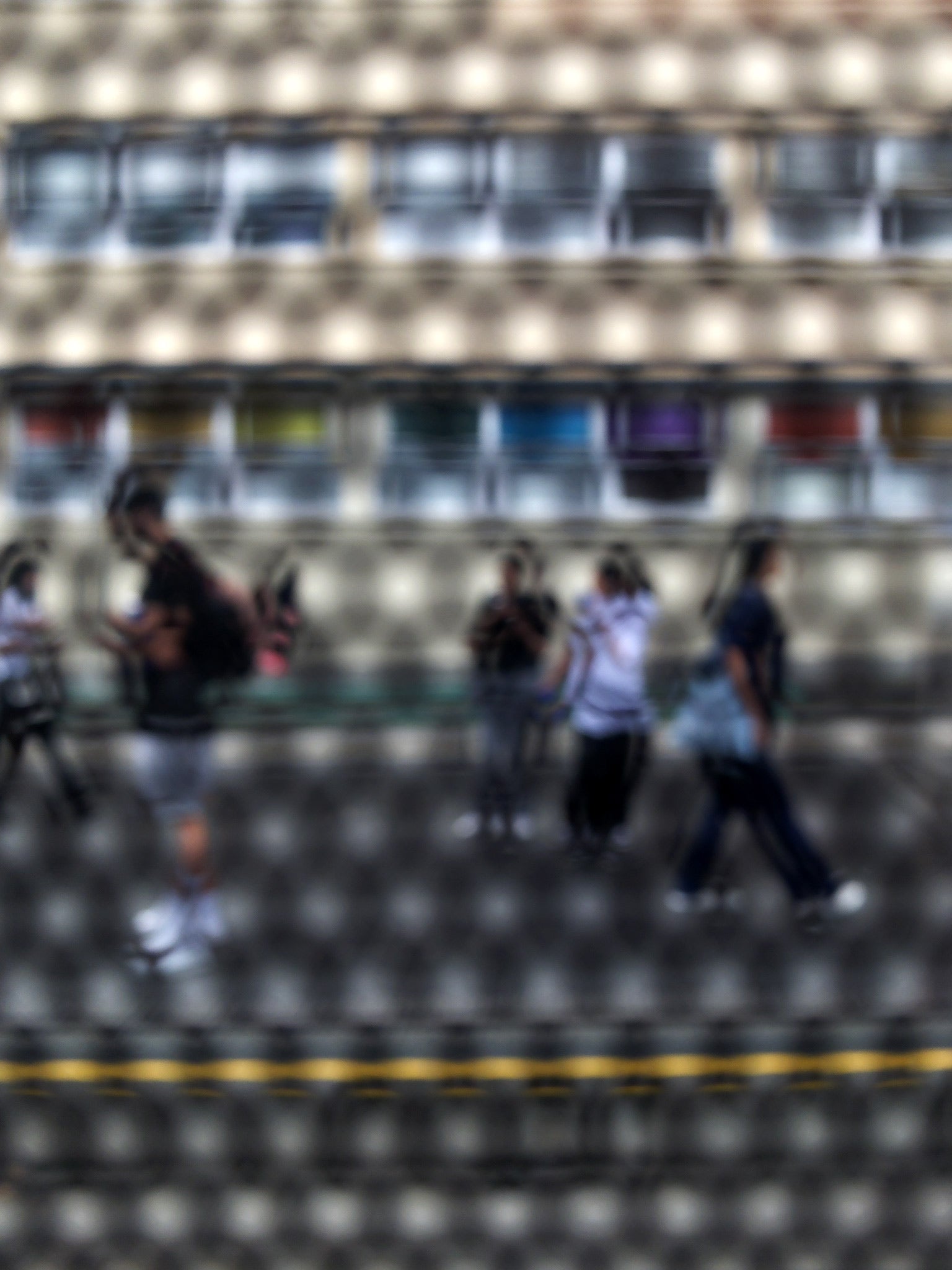 Urban Commute - Series No.2: 9558(1)v1 - 67.5x90cm, fine art photograph by Raymond Mather showing a group of pedestrians walking past a multicoloured glass façade, viewed through a circular screen. The image blurs motion, structure, and rhythm into a poetic urban abstraction.