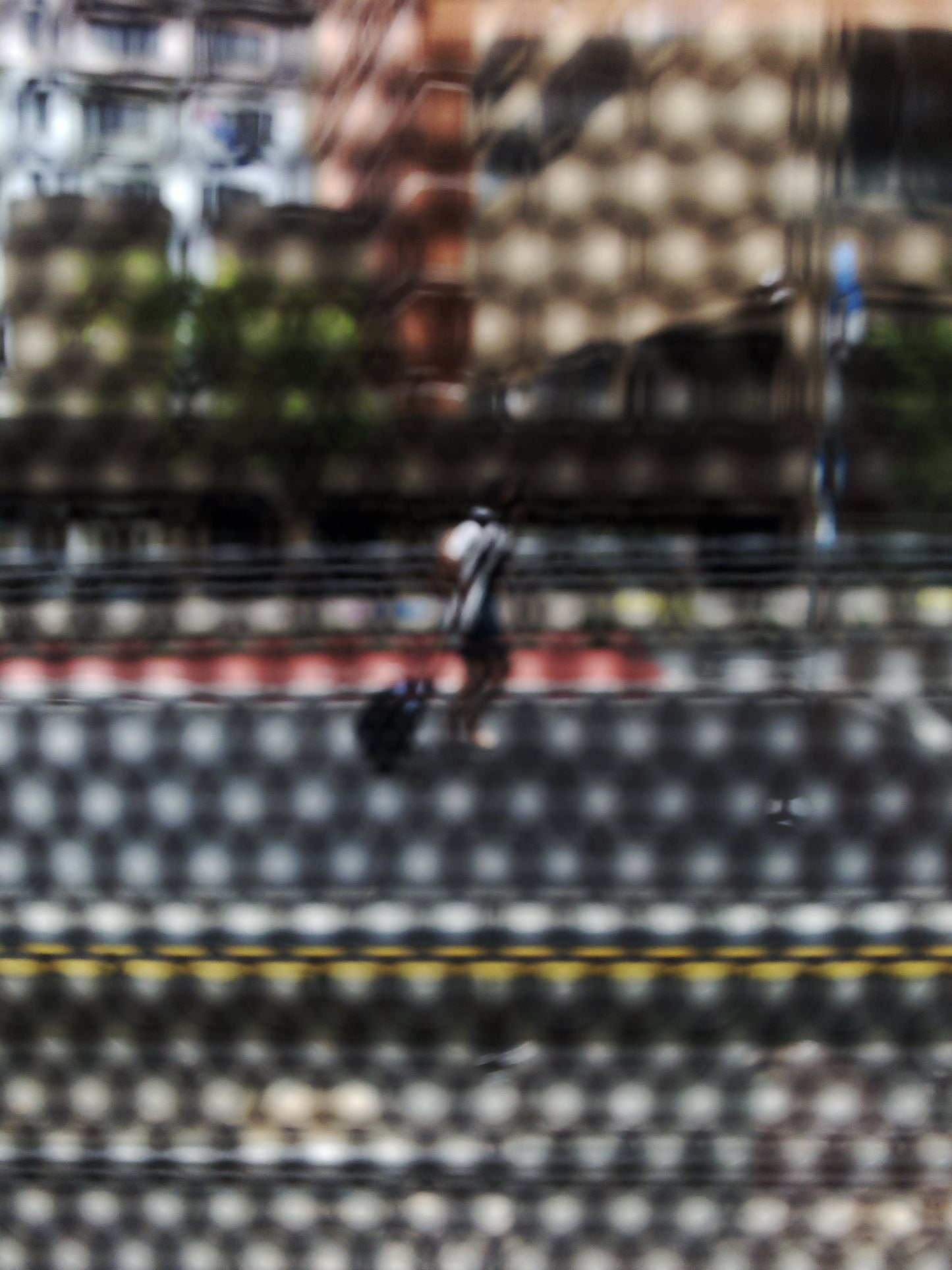 Urban Commute - Series No.2: 9569(1)v1 - 67.5x90cm, fine art photograph by Raymond Mather featuring a lone blurred figure walking along a city street. Viewed through a dotted screen, the image blends red kerb, yellow line, and soft building reflections into a textured urban abstraction.