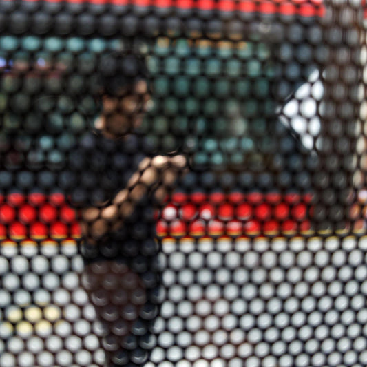Urban Commute - Series No.2: 9574(1)v1 - Close Detail View, fine art photograph by Raymond Mather featuring a blurred figure using a phone, seen through a patterned screen with a red and green bus in the background. A richly layered urban image exploring distraction, movement, and modern life.