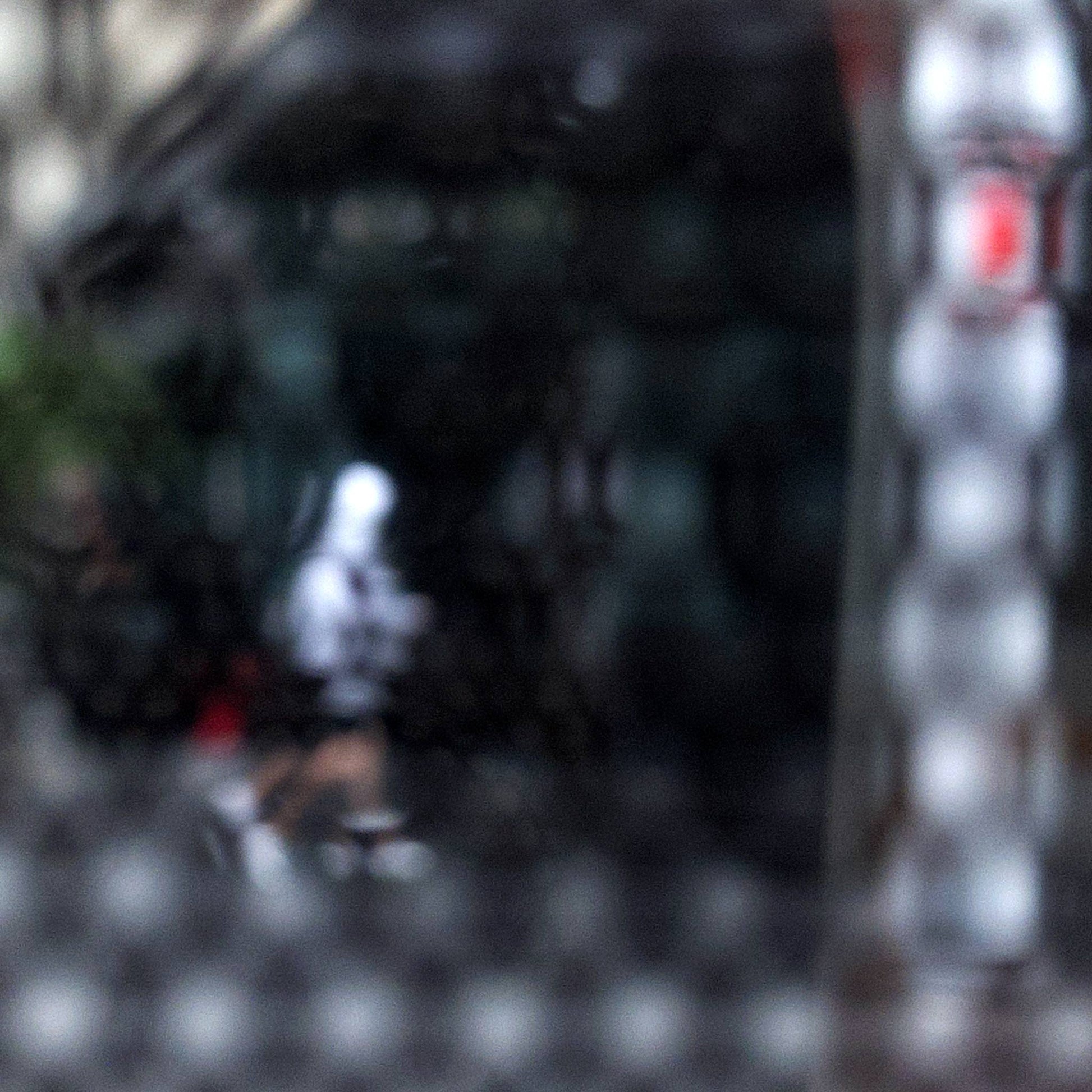 Urban Commute - Series No.2: 9590v1 - 90x90cm, fine art photograph by Raymond Mather showing a blurred white-hooded figure walking across a dark city street, seen through a patterned screen. A haunting study in motion, texture, and introspection.