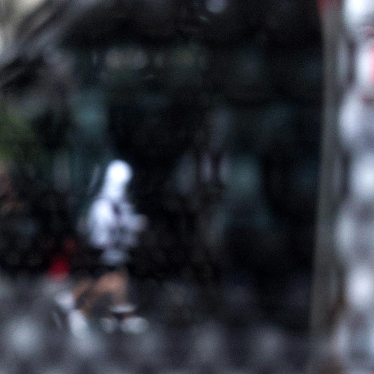 Urban Commute - Series No.2: 9590v1 - Close Detail View, fine art photograph by Raymond Mather showing a blurred white-hooded figure walking across a dark city street, seen through a patterned screen. A haunting study in motion, texture, and introspection.